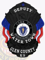 A custom metal badge in black featuring the title "DEPUTY" prominently at the top, above a central eagle emblem. The badge displays the words "COMM. OF MASSACHUSETTS" and "WATER TOWN" in bold letters. Below the state seal, it reads "GLEN COUNTY" and "K9." The badge is adorned with decorative blue and red elements, enhancing its design. The matte black finish gives the badge a sleek and authoritative appearance, emphasizing its significance in law enforcement.