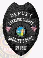 A custom metal badge in black featuring the title "DEPUTY" at the top, followed by "LAKESIDE COUNTY." The center showcases a circular emblem of the state seal of California, flanked by decorative elements including American flags. Below, it reads "SHERIFF'S DEPT." and "K9 UNIT." The matte black finish gives the badge a sleek and authoritative appearance, emphasizing its significance in law enforcement and the K9 unit's role in the community.