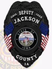 A custom metal badge in black featuring the title "DEPUTY" at the top, along with the name "JACKSON" and "SINCE 1905." The center showcases a circular emblem of the state of Iowa, flanked by decorative blue panels and American flags on either side. Below the emblem, the badge reads "COUNTY IA." The matte black finish gives the badge a sleek and modern appearance, emphasizing its official status and authority in law enforcement.